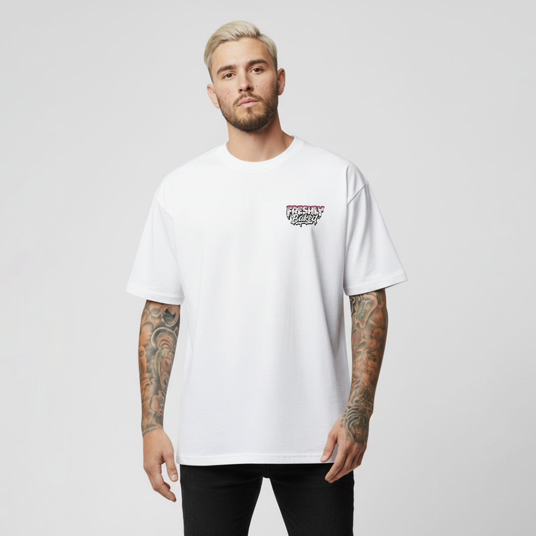 A man with blonde hair and tattoos wearing a white short-sleeve t-shirt with a small graphic on the left chest, paired with black pants, against a light gray background.