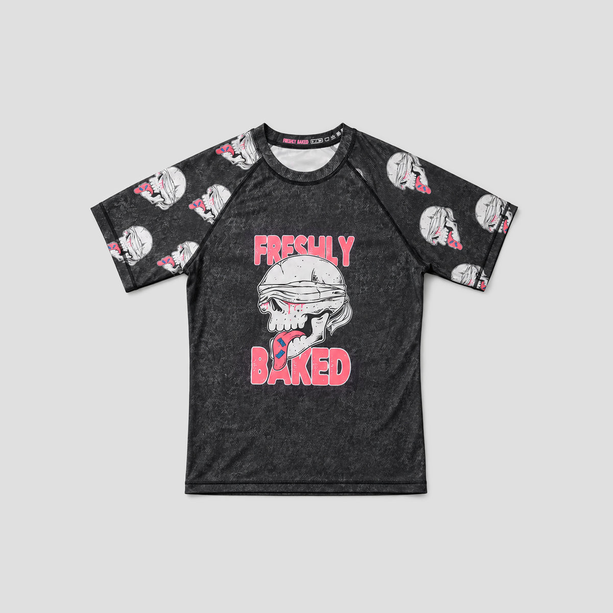 A short-sleeve raglan t-shirt in a dark charcoal gray mineral wash, featuring a graphic of a blindfolded skull with pink text reading 'FRESHLY BAKED' on the front. The sleeves and collar area feature a repeating pattern of white skulls wearing pink bow ties.