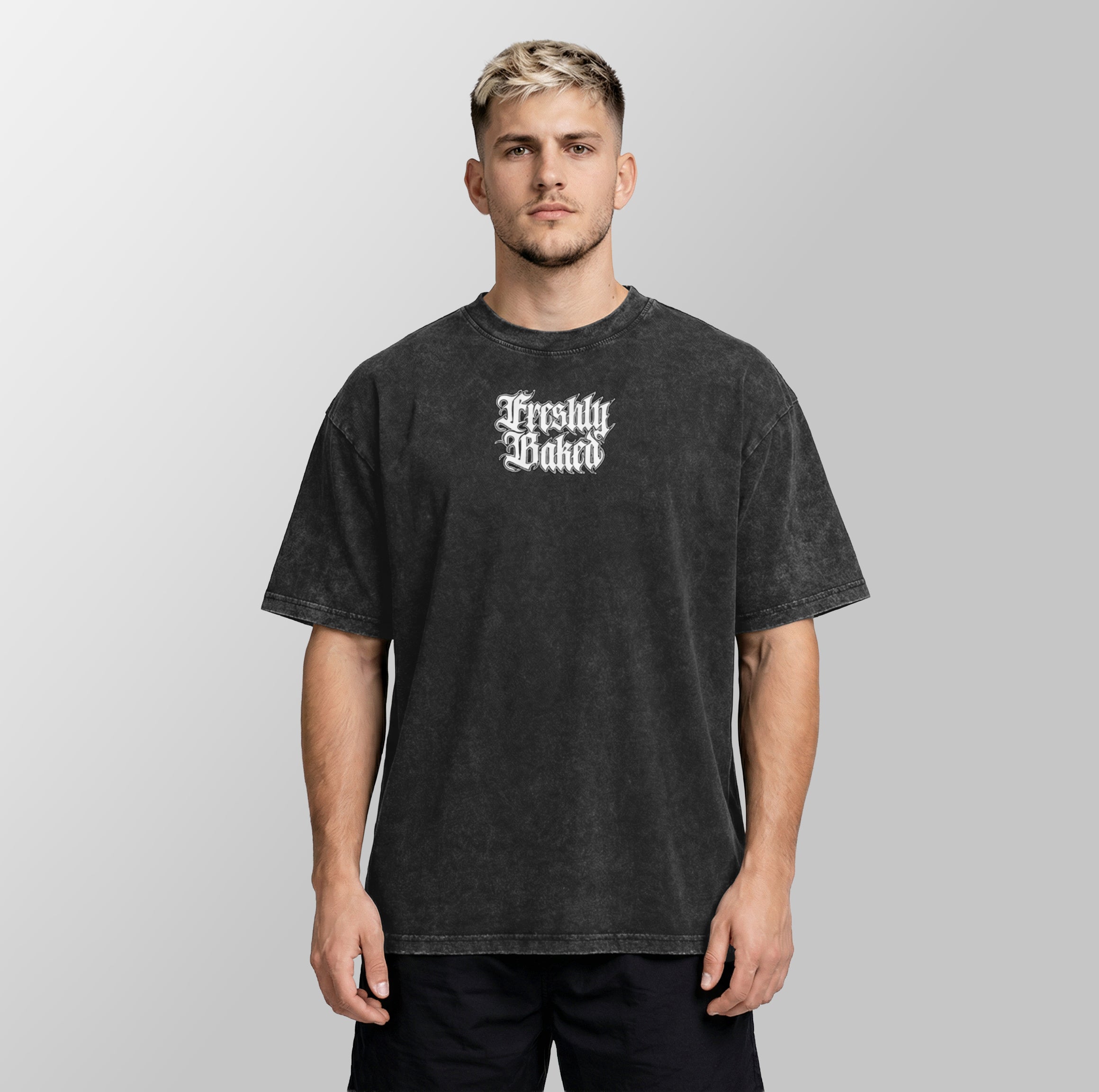 A man wearing a faded black graphic t-shirt with 'Freshly Baked' printed in white Gothic lettering on the chest, set against a plain light gray background.