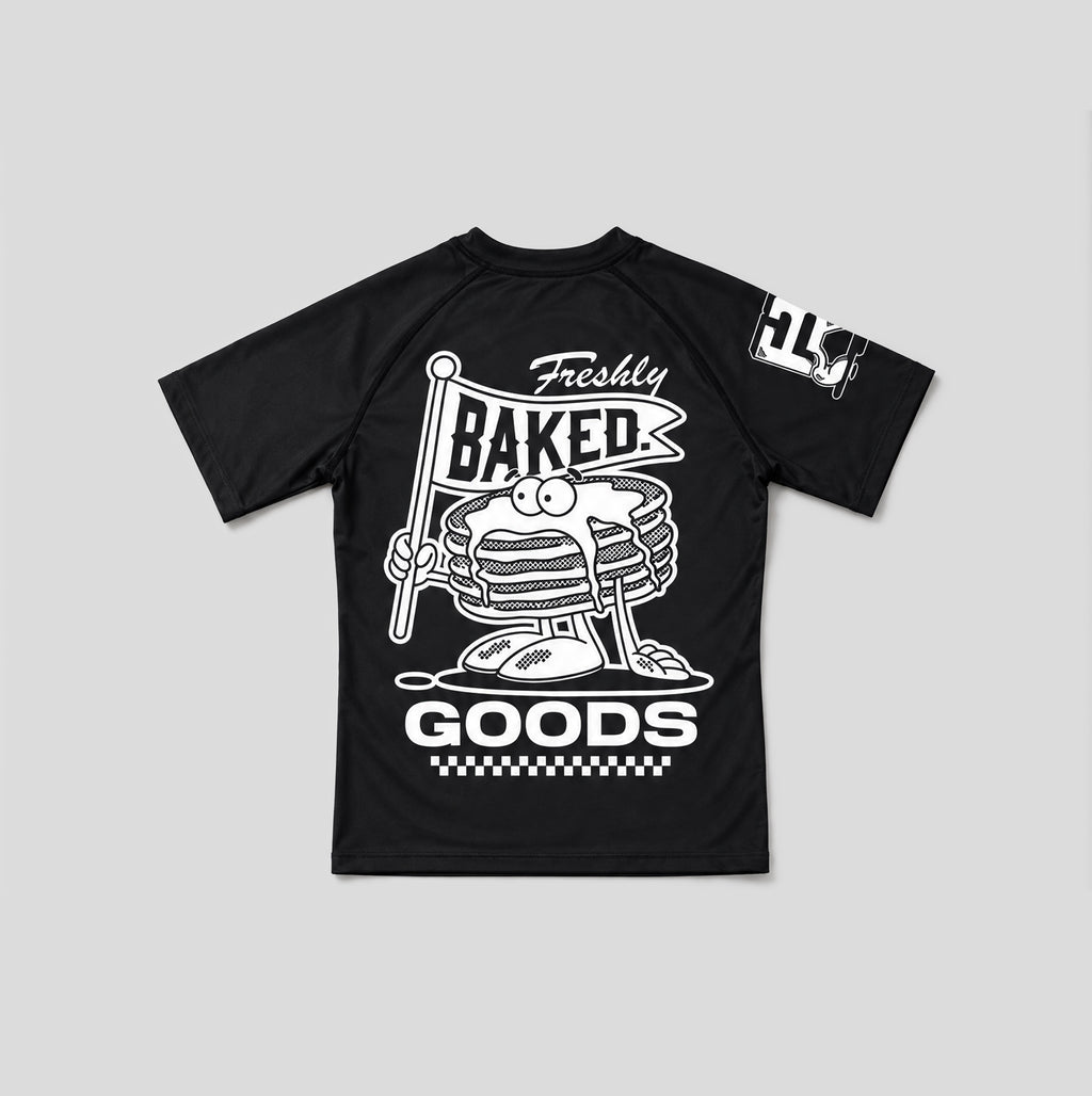 A black short-sleeve raglan t-shirt with a large white graphic on the front featuring a cartoon stack of pancakes holding a 'Freshly Baked' sign, and a white graphic on the right sleeve.