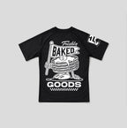 A black short-sleeve raglan t-shirt with a large white graphic on the front featuring a cartoon stack of pancakes holding a 'Freshly Baked' sign, and a white graphic on the right sleeve.