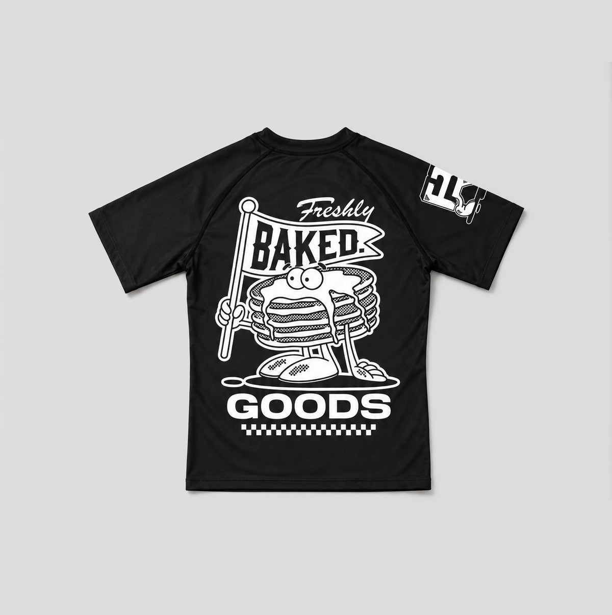 A black short-sleeve raglan t-shirt with a large white graphic on the front featuring a cartoon stack of pancakes holding a 'Freshly Baked' sign, and a white graphic on the right sleeve.