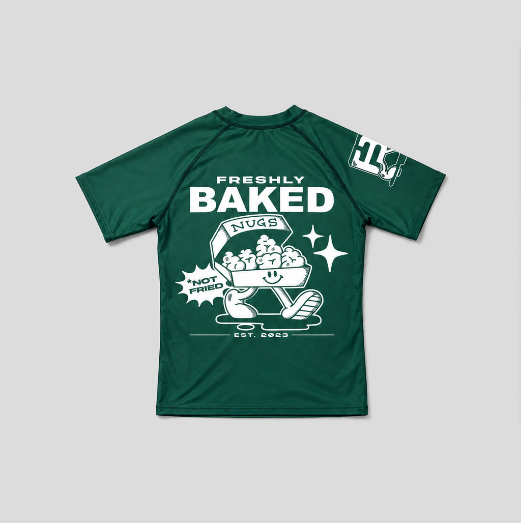 A dark green short-sleeve t-shirt with a white graphic print on the front reading 'FRESHLY BAKED NUGS' and 'HOT DUTCH', alongside a cartoon bagel character. A smaller white graphic is visible on the right sleeve.