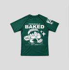 A dark green short-sleeve t-shirt with a white graphic print on the front reading 'FRESHLY BAKED NUGS' and 'HOT DUTCH', alongside a cartoon bagel character. A smaller white graphic is visible on the right sleeve.
