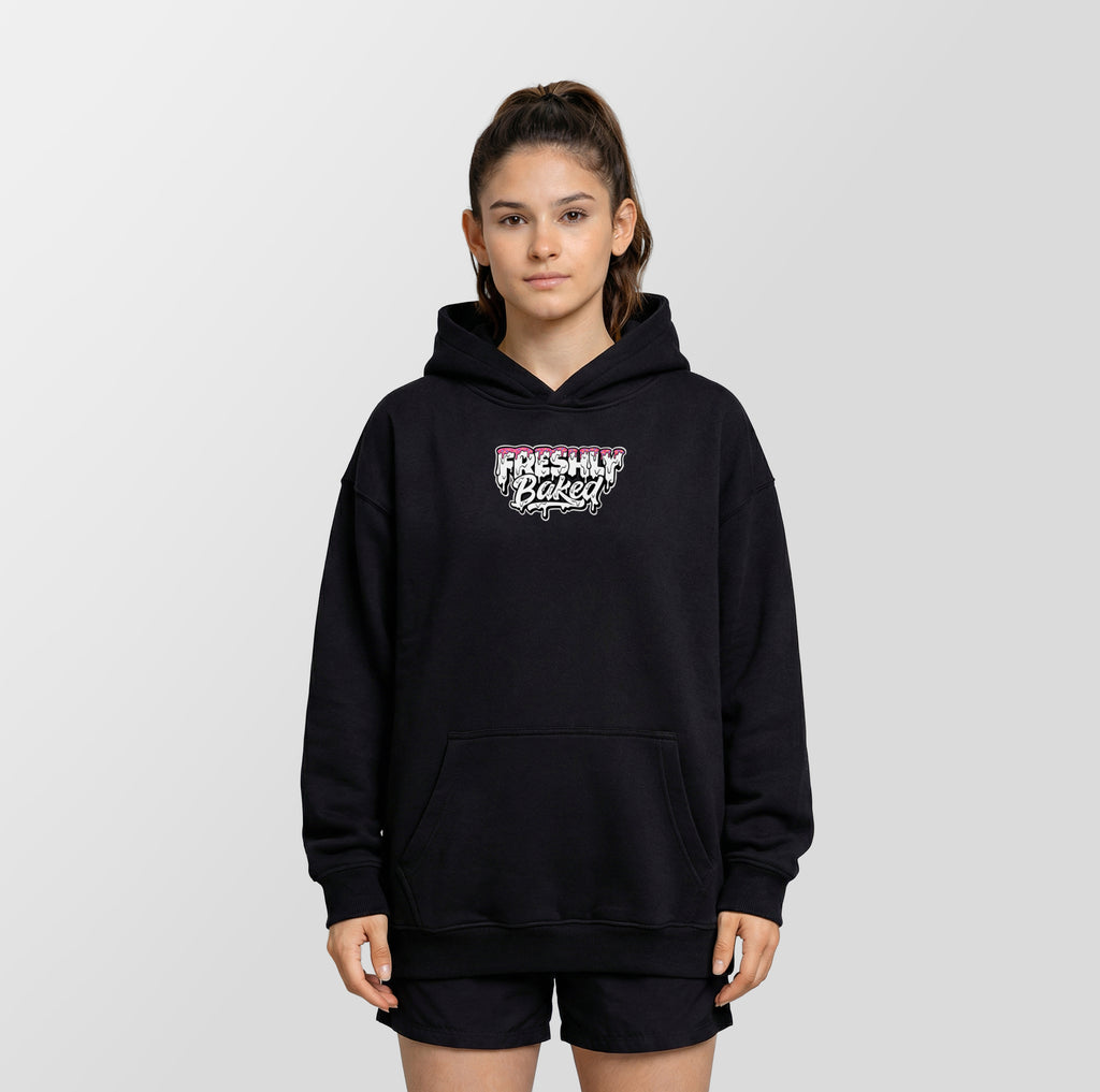 A young woman modeling a black pullover hoodie with a 'Freshly Baked' graphic on the chest, paired with black athletic shorts, against a light gray studio background.