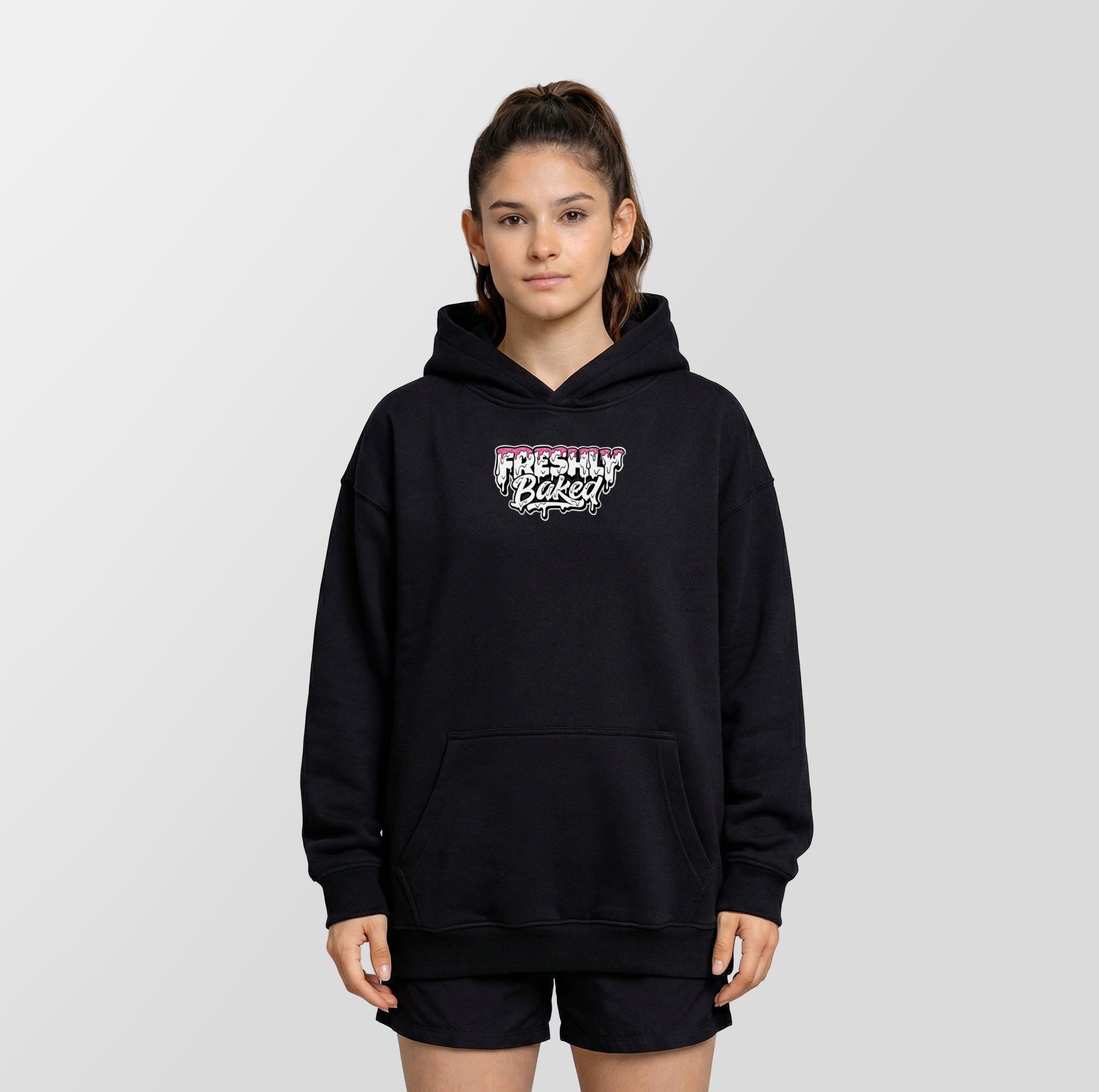A young woman modeling a black pullover hoodie with a 'Freshly Baked' graphic on the chest, paired with black athletic shorts, against a light gray studio background.