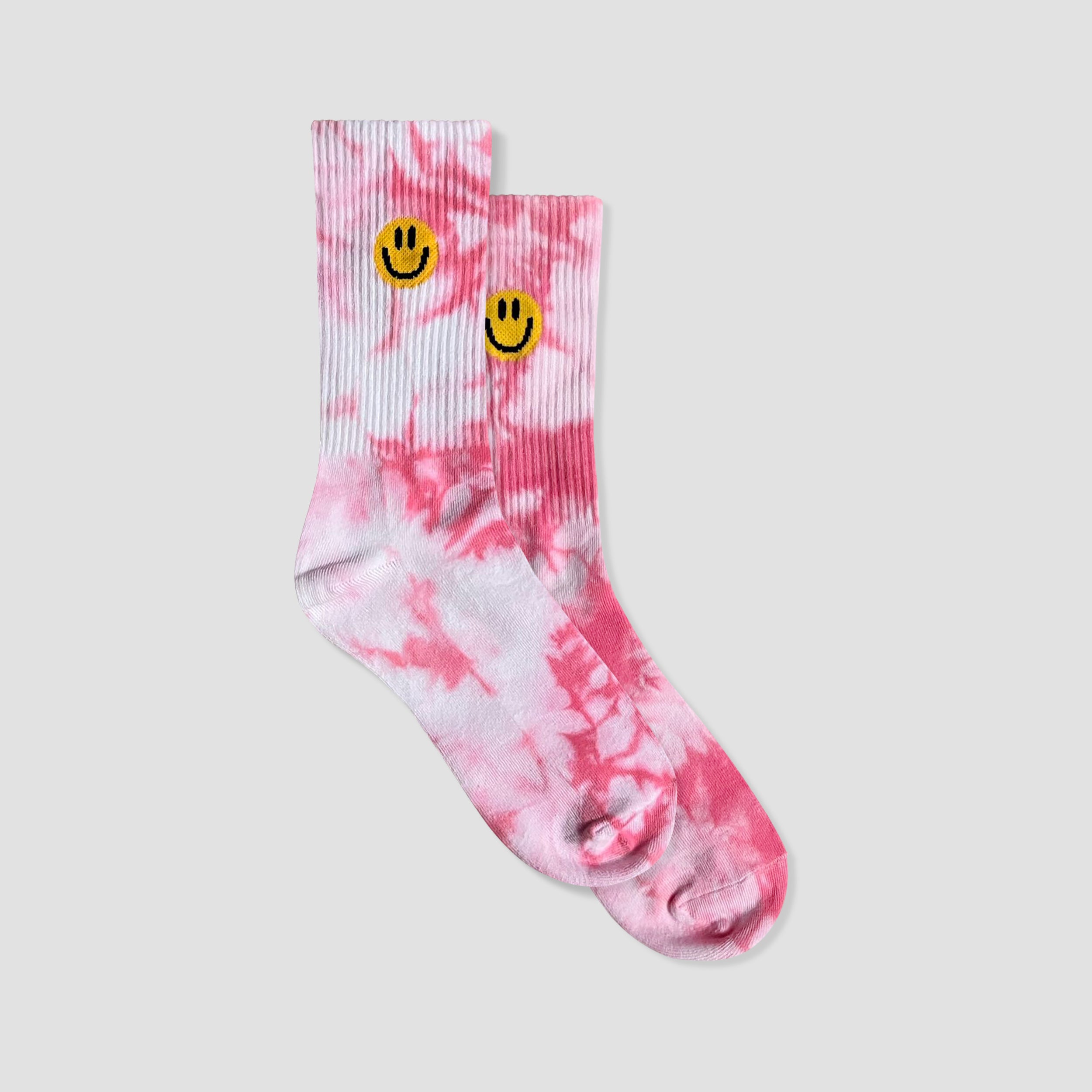 A pair of pink and white tie-dye crew socks with yellow smiley face graphics on the ribbed cuffs, displayed against a solid light gray background.