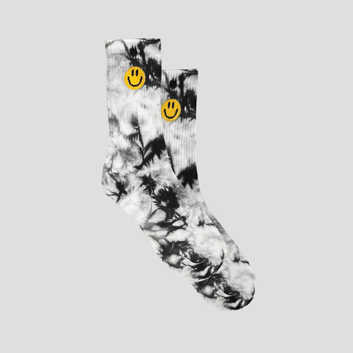 A pair of crew socks featuring a black and white tie-dye pattern with a yellow smiley face printed near the cuff, displayed against a solid light gray background.