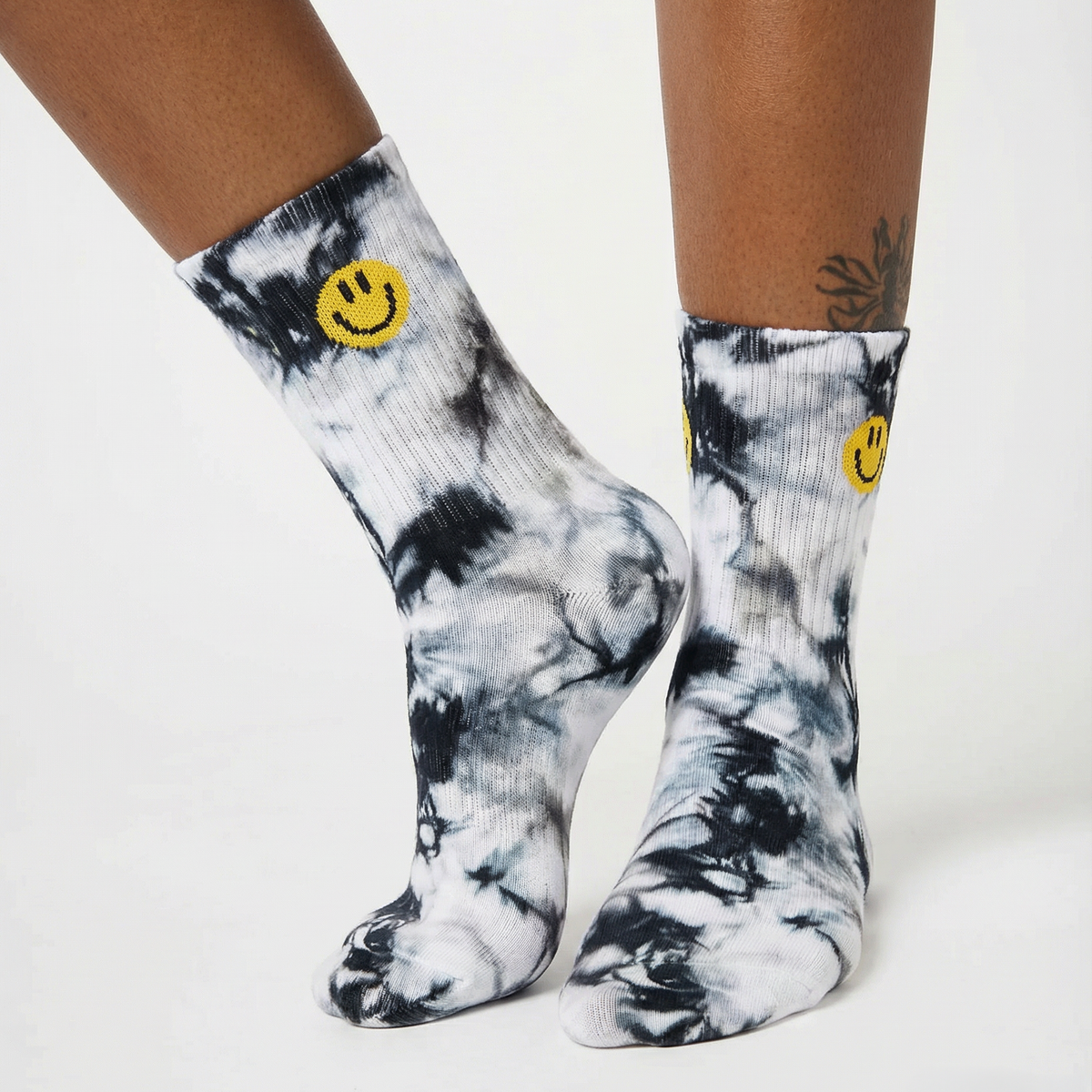 A pair of crew socks with a black and white tie-dye pattern and a yellow smiley face logo near the cuff, shown on a model's feet against a white studio background.