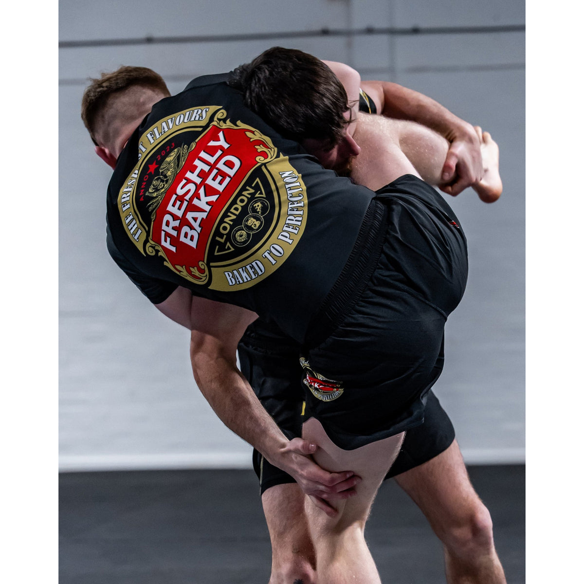 Two people grappling in a gym, with the person on top wearing black shorts and a black top featuring a large red, gold, and white circular graphic on the back that reads 'Freshly Baked London'.