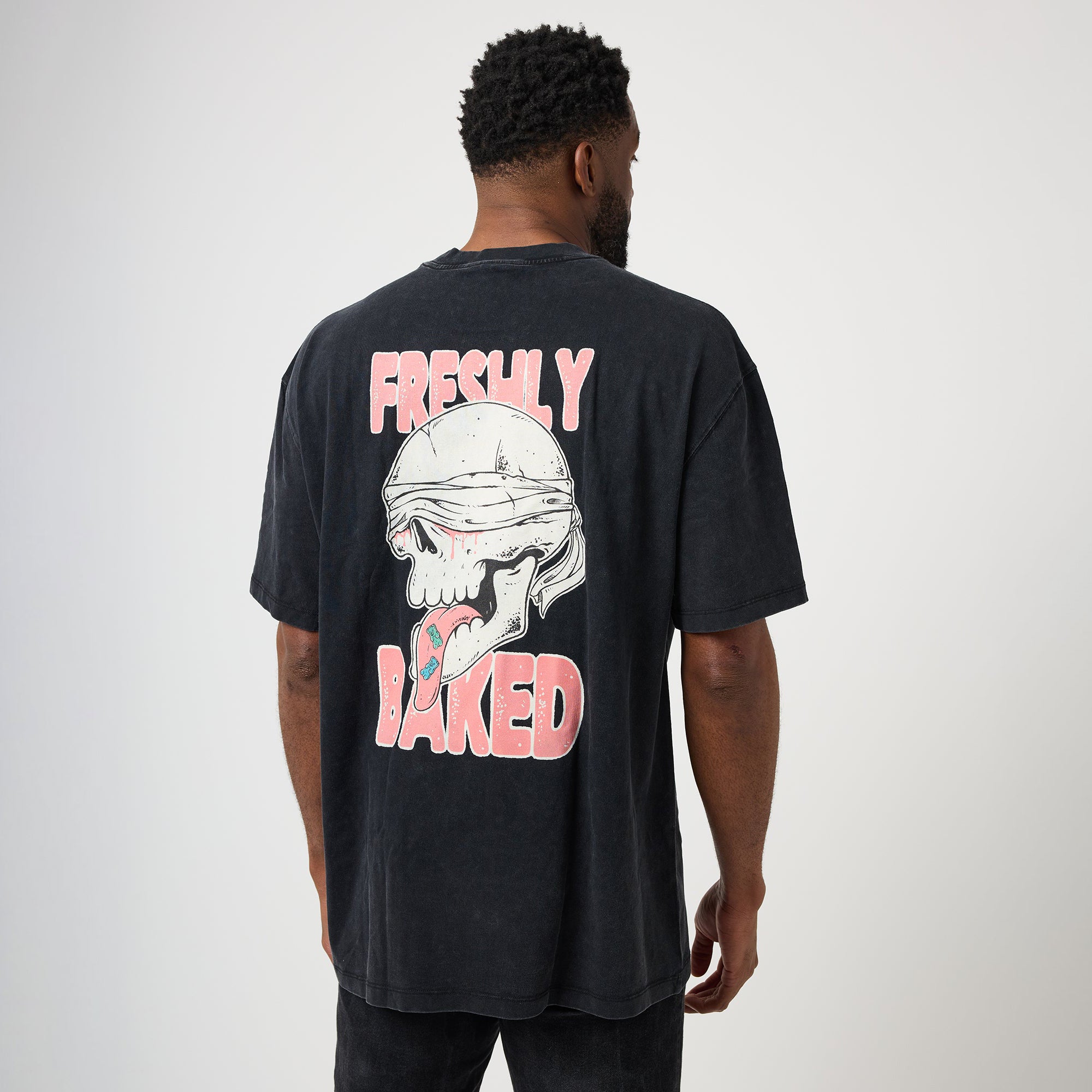 A man viewed from behind wearing a faded black t-shirt with a large graphic on the back featuring a skull and the words 'FRESHLY BAKED' in pink and white.