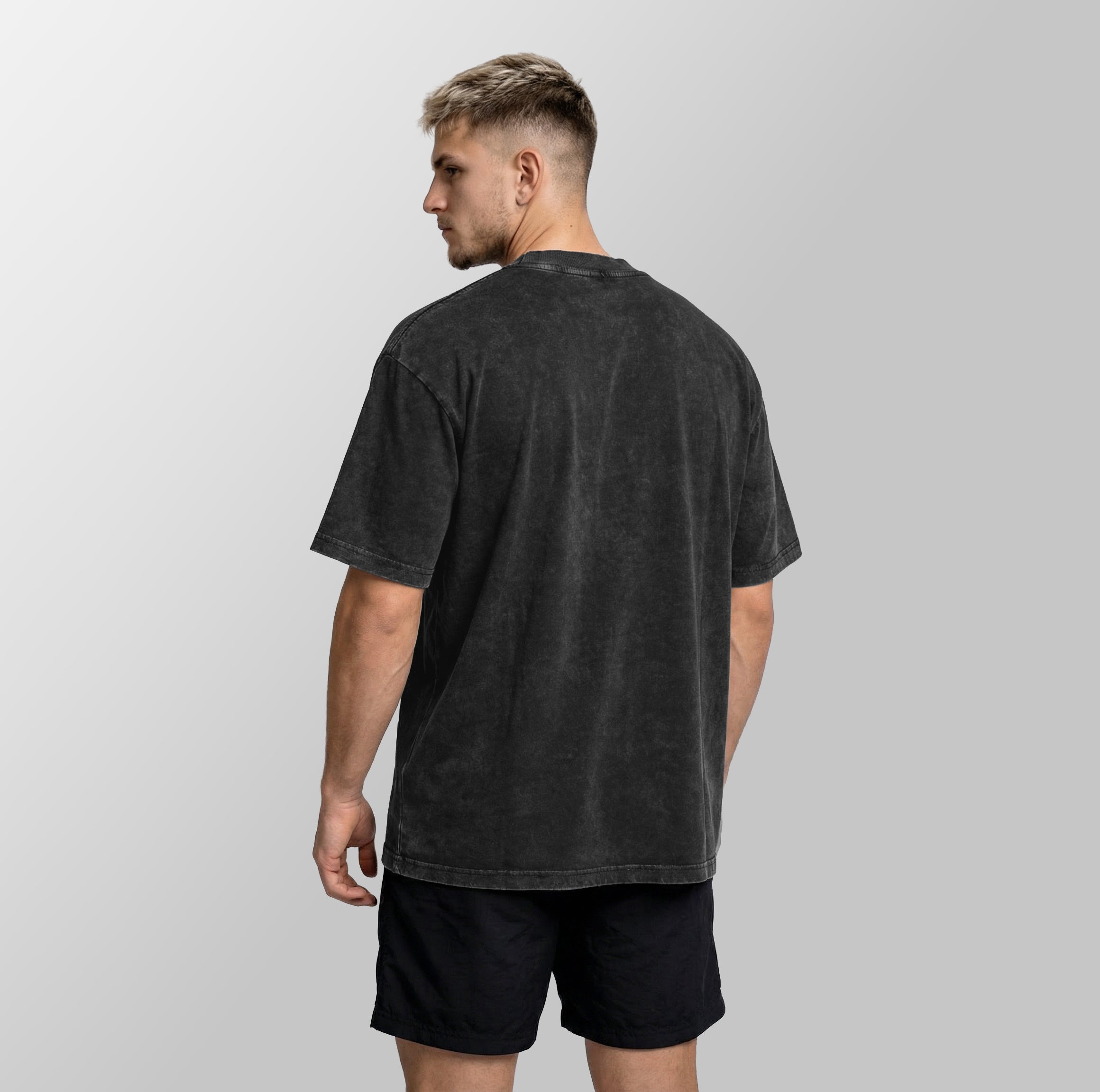 A man wearing a faded black acid-wash t-shirt and black shorts, shown from the back against a light gray studio background.