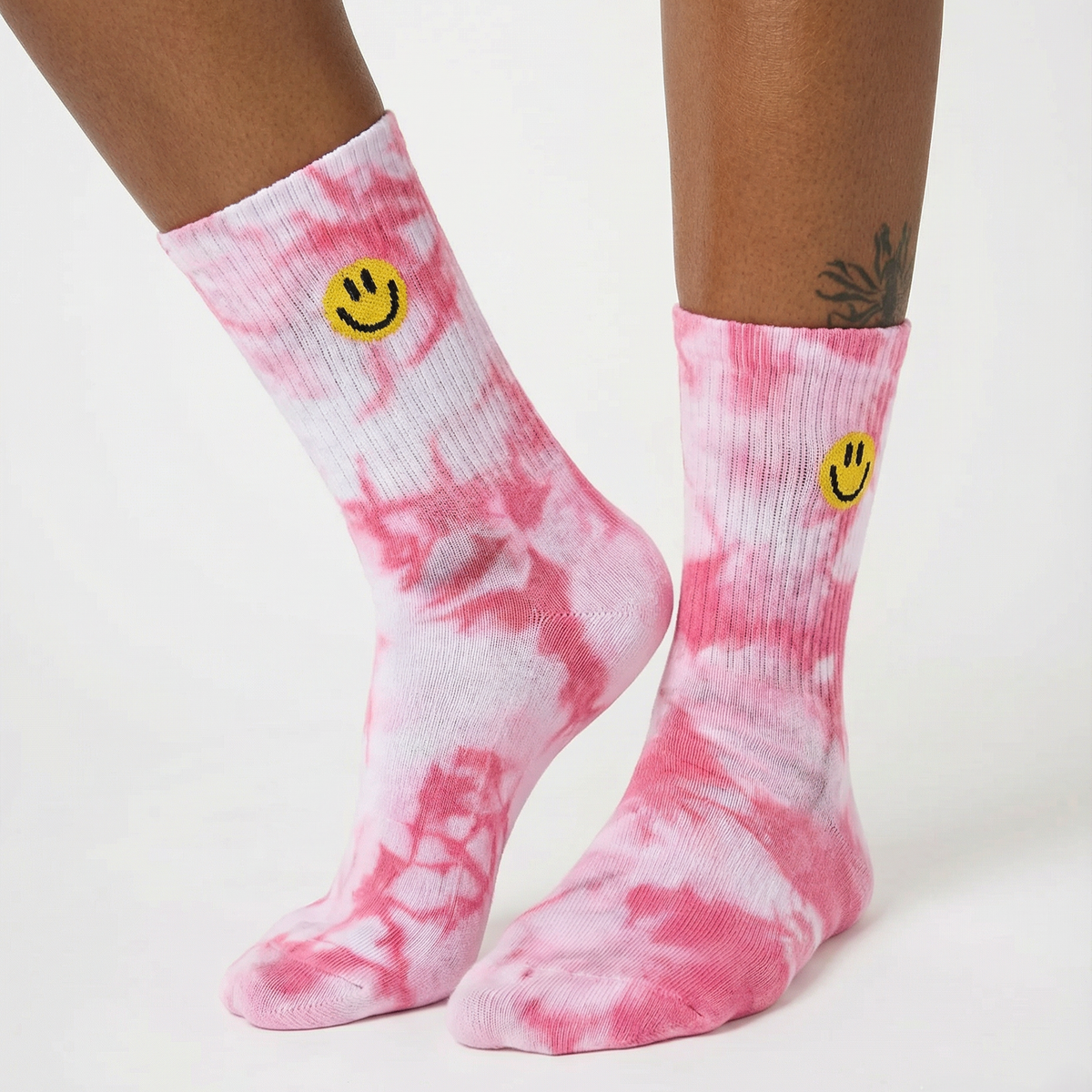Pink and white tie-dye crew socks with a yellow smiley face logo near the cuff, shown on a model's feet against a plain white background.