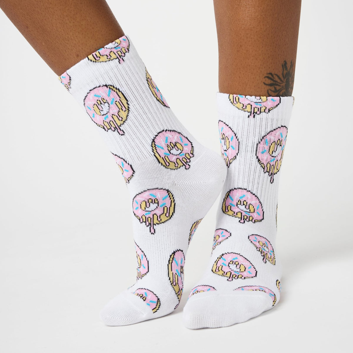White crew socks with a repeating pattern of dripping donuts in pink, yellow, and blue, shown on a model's feet against a light gray studio background.
