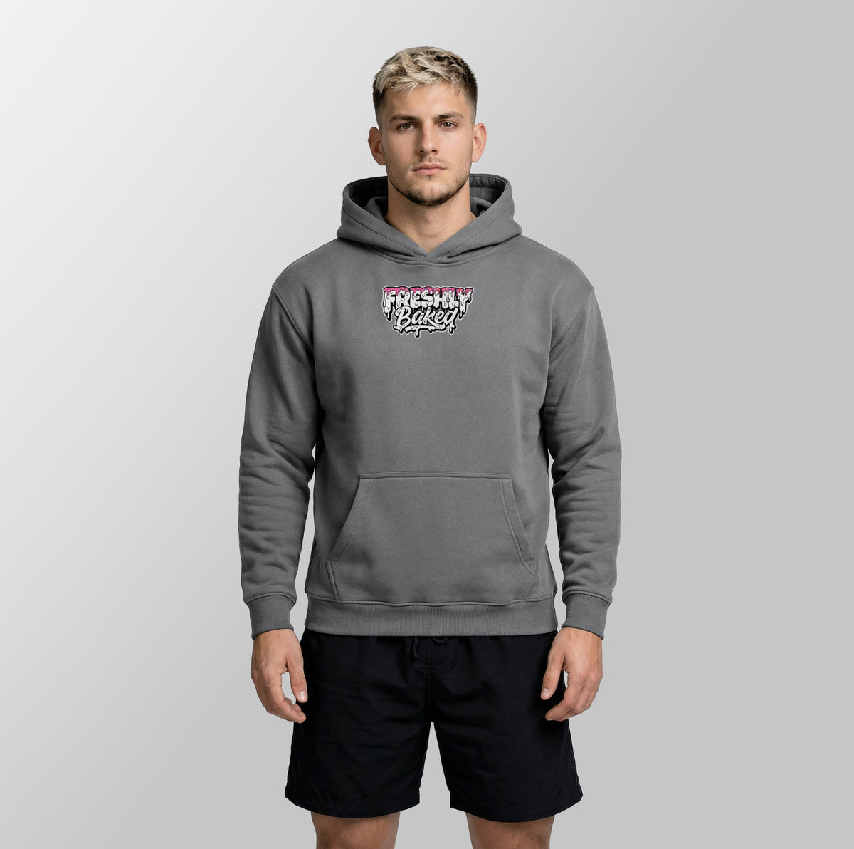 A man wearing a gray pullover hoodie with a 'Freshly Baked' graphic on the chest, paired with black athletic shorts, standing against a light gray studio background.