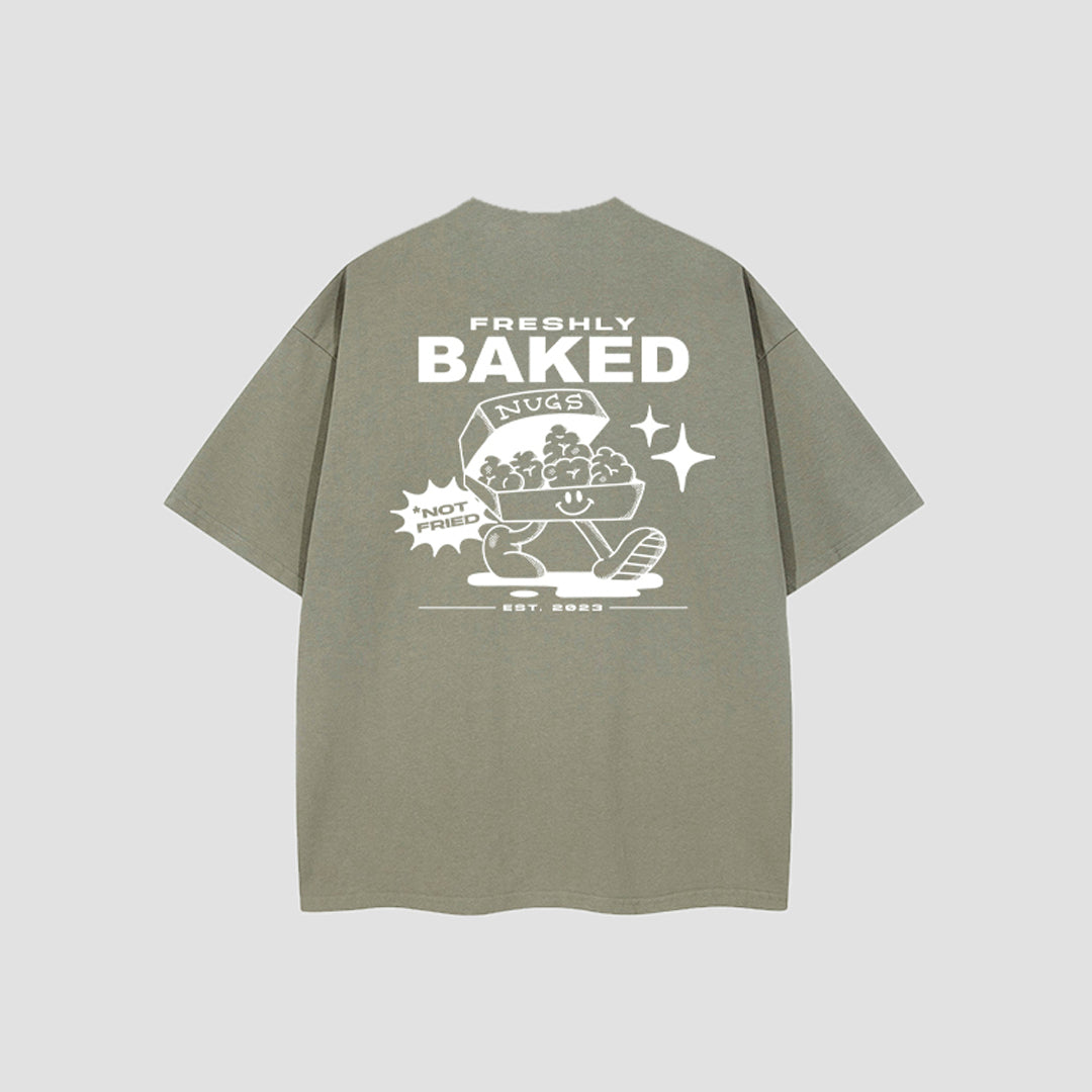 An oversized short-sleeve t-shirt in a muted sage green color, shown from the back. It features a large white graphic print with the words 'FRESHLY BAKED', a bag of chips, and a foot walking out of a loaf of bread, with 'EST.