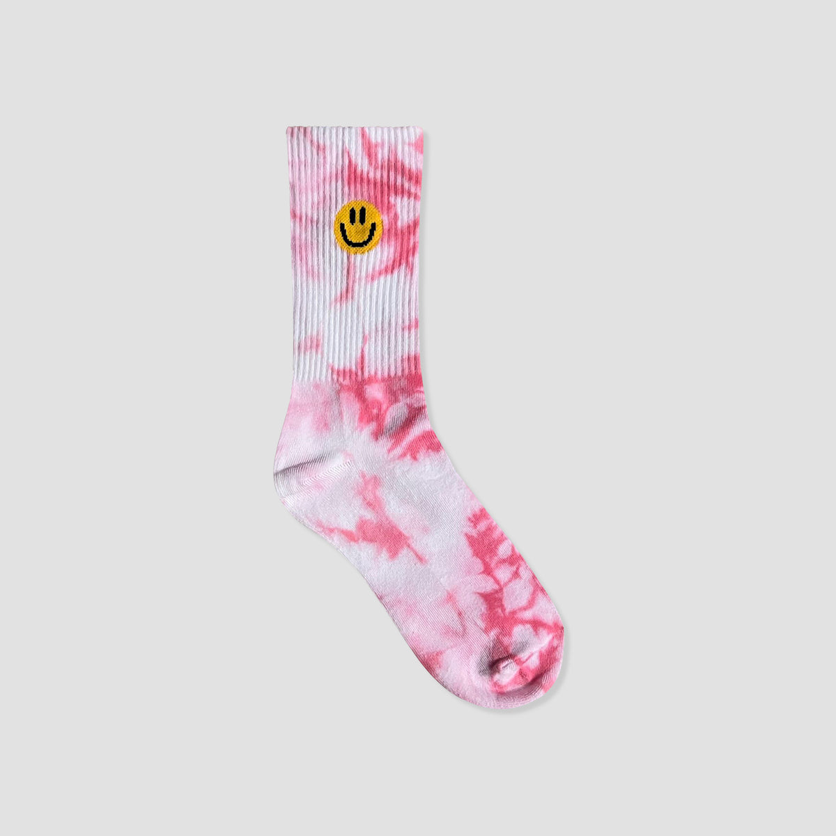A single pink and white tie-dye crew sock with a yellow smiley face embroidered on the ribbed cuff, shown against a solid light gray background.
