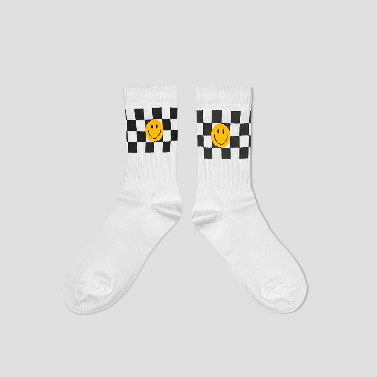 A pair of white crew socks with a black and white checkered pattern and a yellow smiley face graphic on the upper calf, displayed against a solid light gray background.