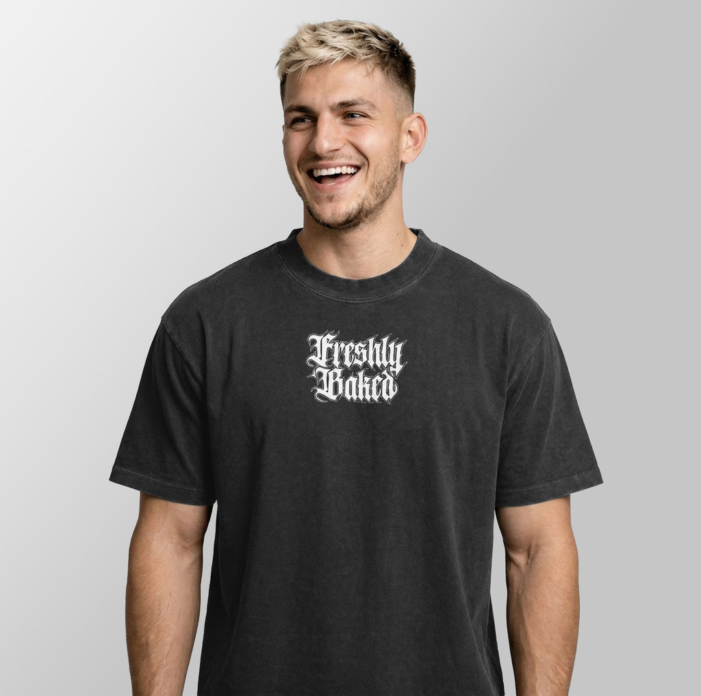 A man smiling and wearing a dark charcoal gray t-shirt with a white gothic-style 'Freshly Baked' graphic on the chest, set against a plain light gray background.