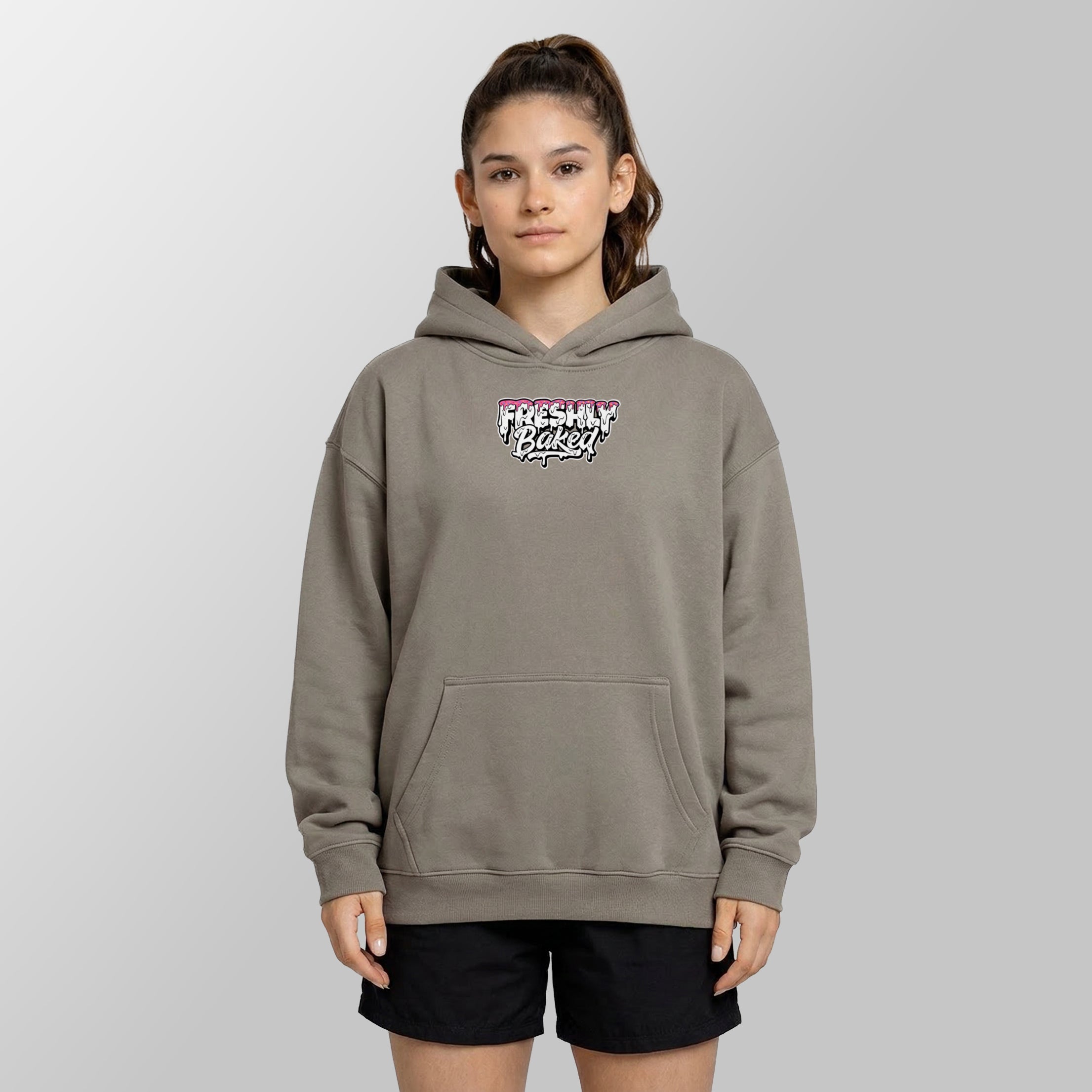 A woman modeling a taupe gray pullover hoodie with a 'Freshly Baked!' graphic on the chest, paired with black shorts, against a neutral gray background.