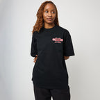 A woman with curly light brown hair wearing a black short-sleeve t-shirt with a red and white graphic on the left chest, paired with black pants, standing against a light gray studio background.