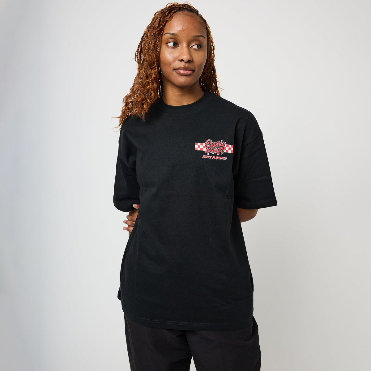 A woman with curly light brown hair wearing a black short-sleeve t-shirt with a red and white graphic on the left chest, paired with black pants, standing against a light gray studio background.