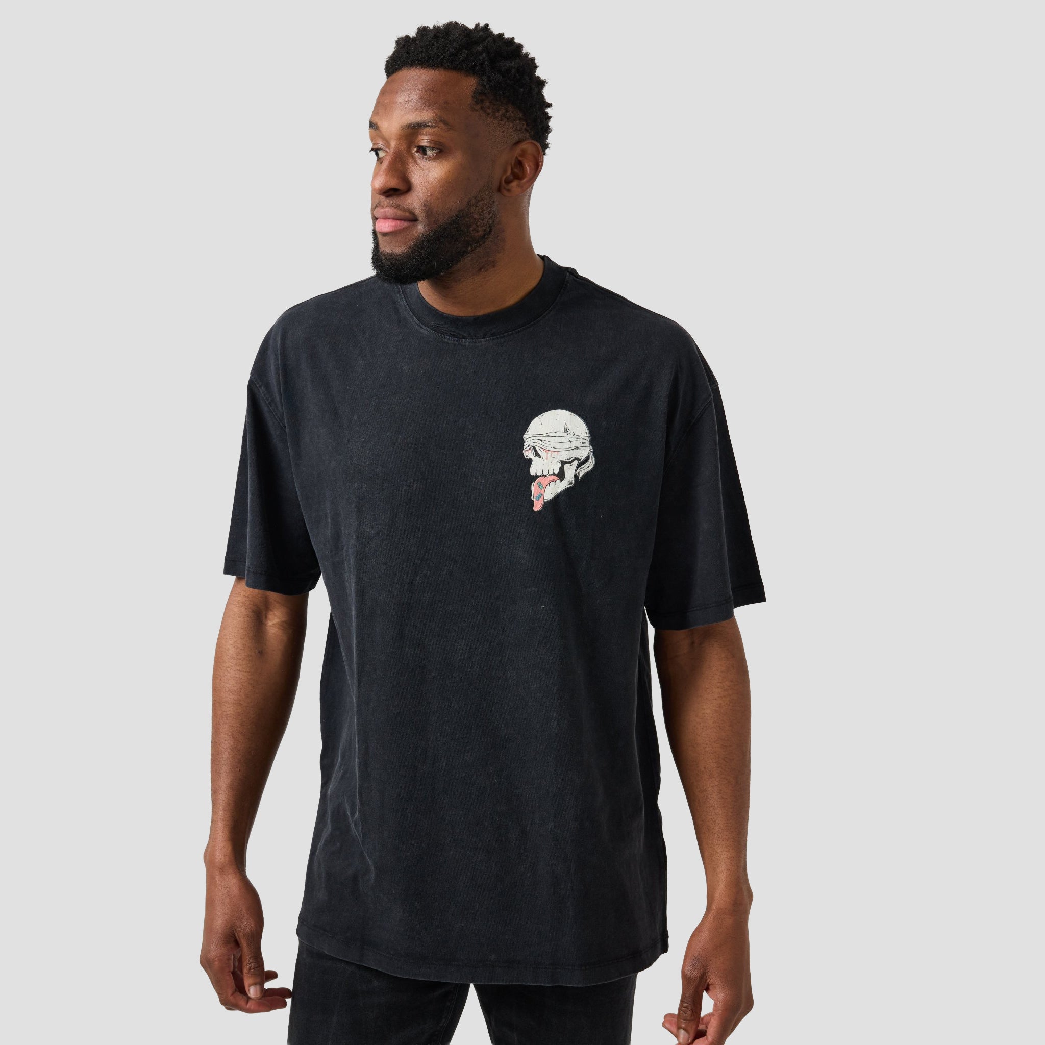 A man wearing a faded black t-shirt with a small graphic of a skull wearing a helmet on the left chest, photographed against a solid light gray background.