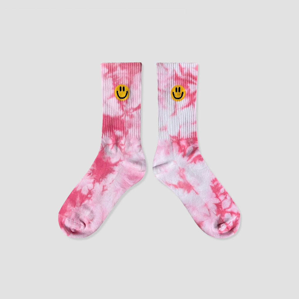 A pair of pink and white tie-dye crew socks with a small yellow smiley face embroidered on the ribbed cuffs, displayed against a solid light gray background.
