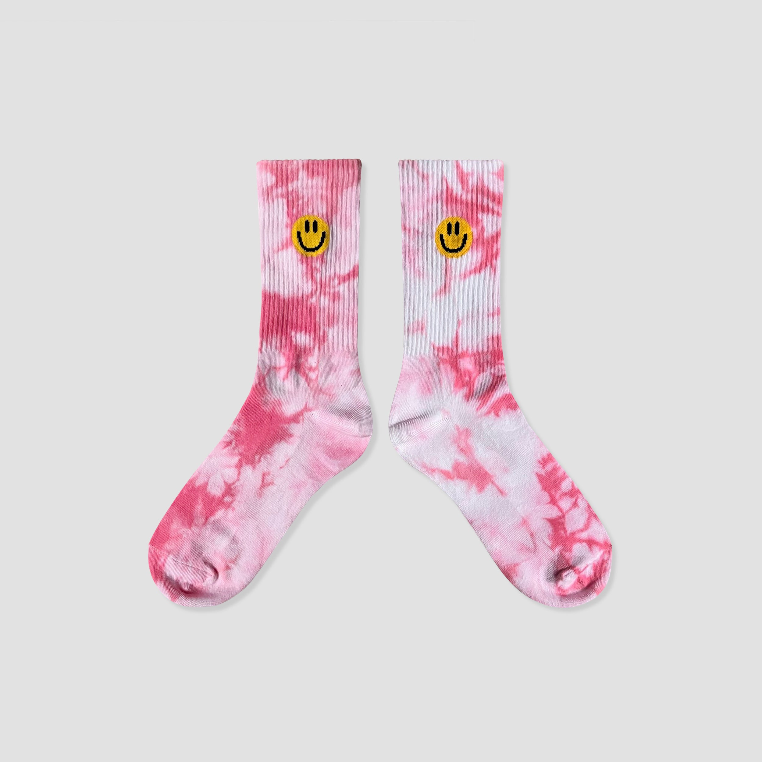 A pair of pink and white tie-dye crew socks with a small yellow smiley face embroidered on the ribbed cuffs, displayed against a solid light gray background.