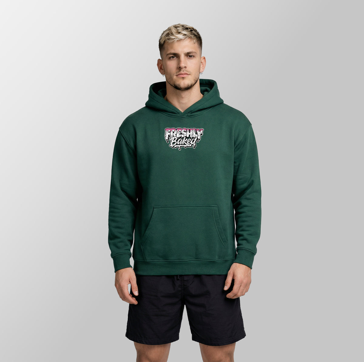 A man wearing a dark green pullover hoodie with a 'Freshly Baked' graphic on the chest, paired with black athletic shorts, standing against a light gray studio background.
