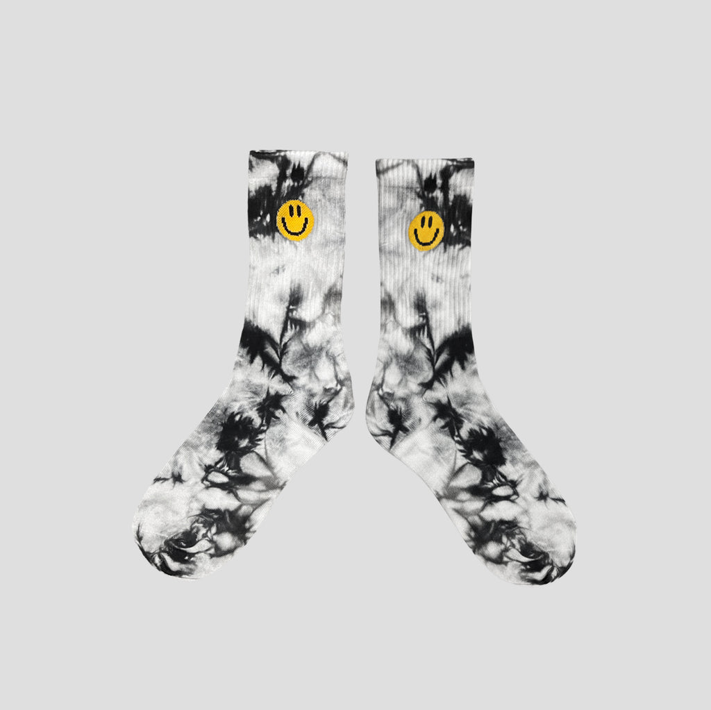 A pair of crew socks with a black and white tie-dye pattern, featuring a small yellow smiley face logo on the ribbed cuffs, displayed against a solid light gray background.