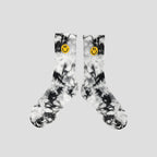 A pair of crew socks with a black and white tie-dye pattern, featuring a small yellow smiley face logo on the ribbed cuffs, displayed against a solid light gray background.