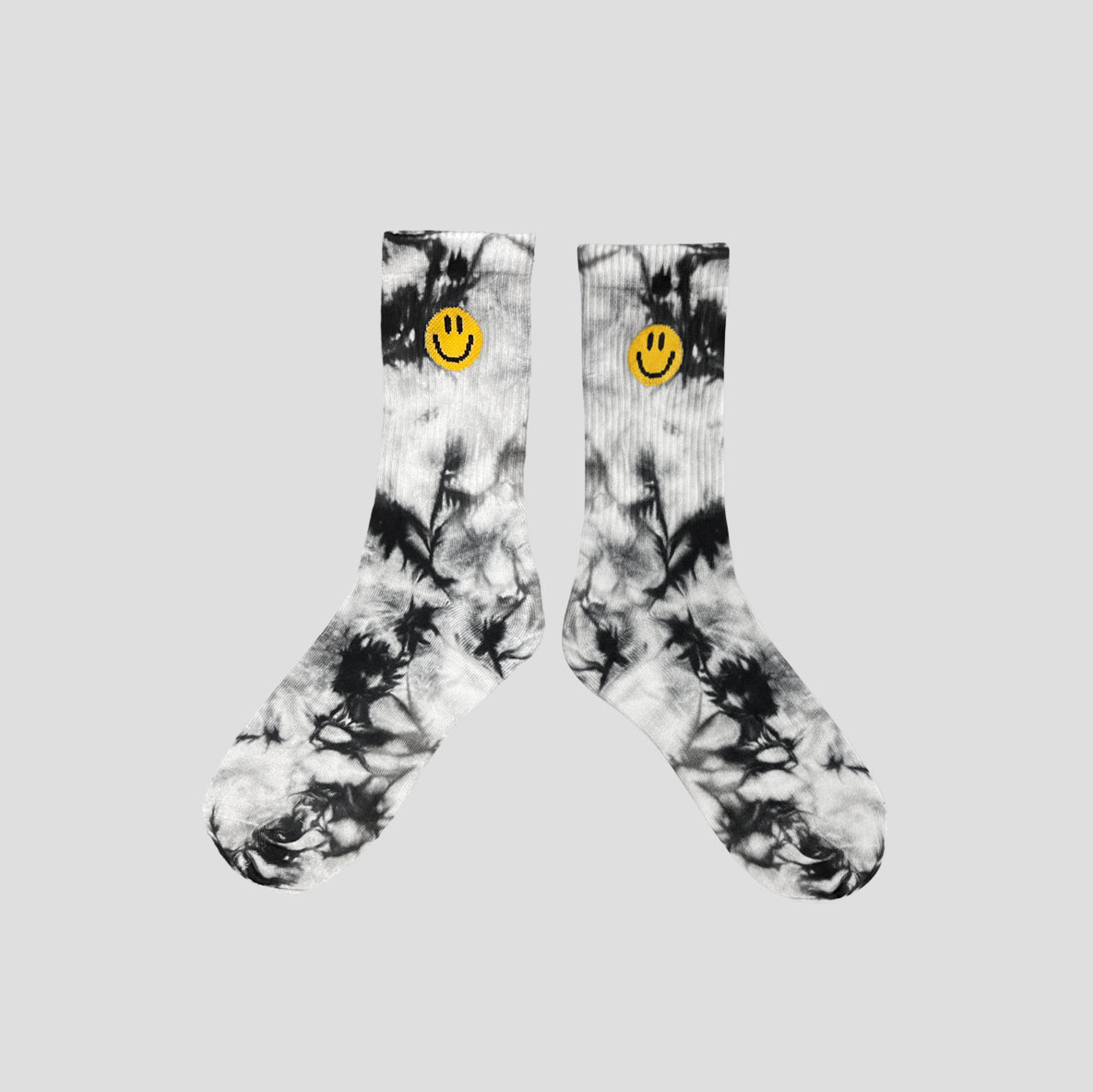 A pair of crew socks with a black and white tie-dye pattern, featuring a small yellow smiley face logo on the ribbed cuffs, displayed against a solid light gray background.