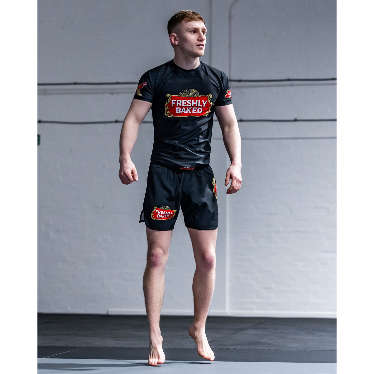 A man wearing a black athletic rash guard and matching black shorts with red and gold 'Freshly Baked' logos, standing barefoot on a mat in front of a white wall.