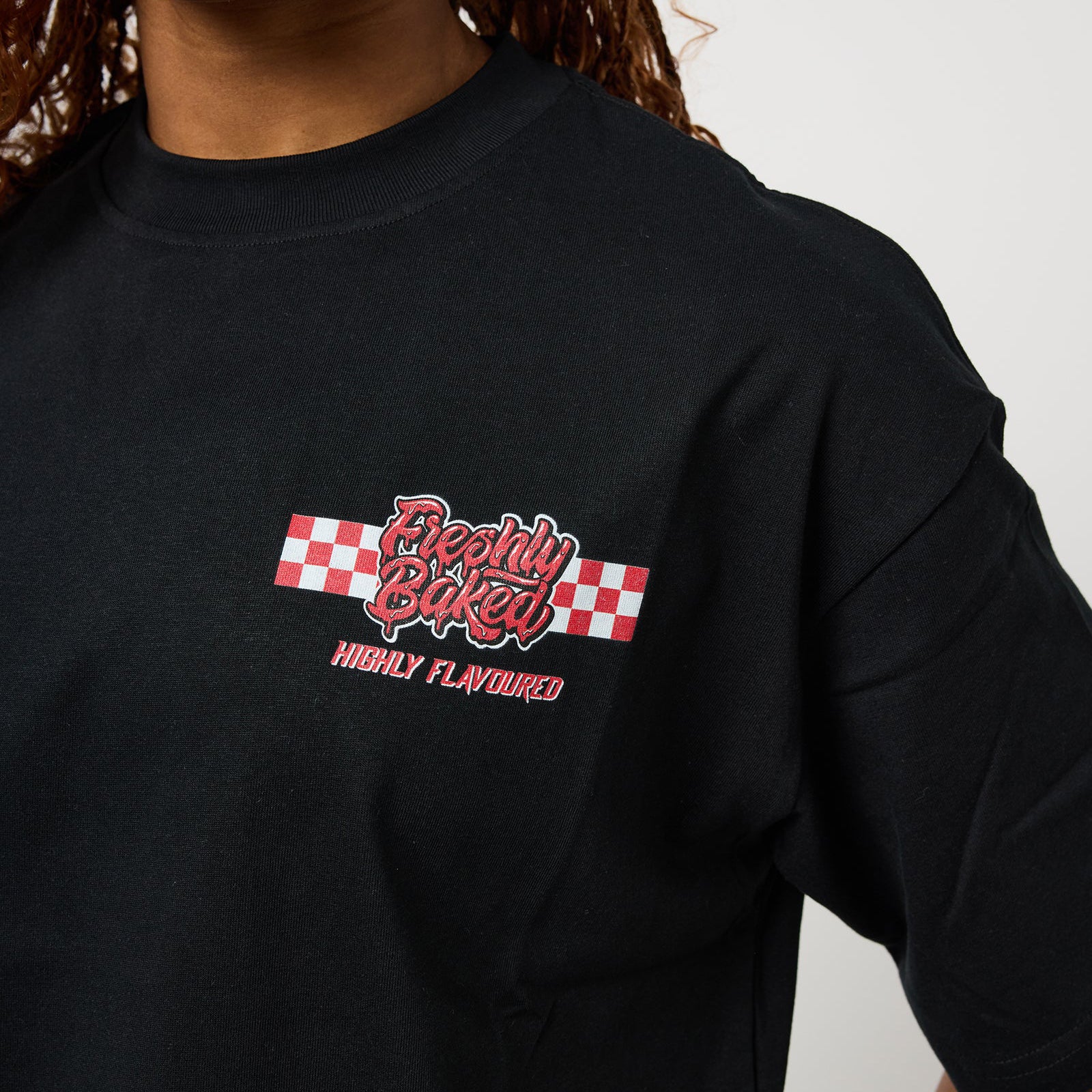 A close-up of a person wearing a black t-shirt with a red and white graphic on the chest that reads 'Freshly Baked' and 'HIGHLY FLAVOURED'.