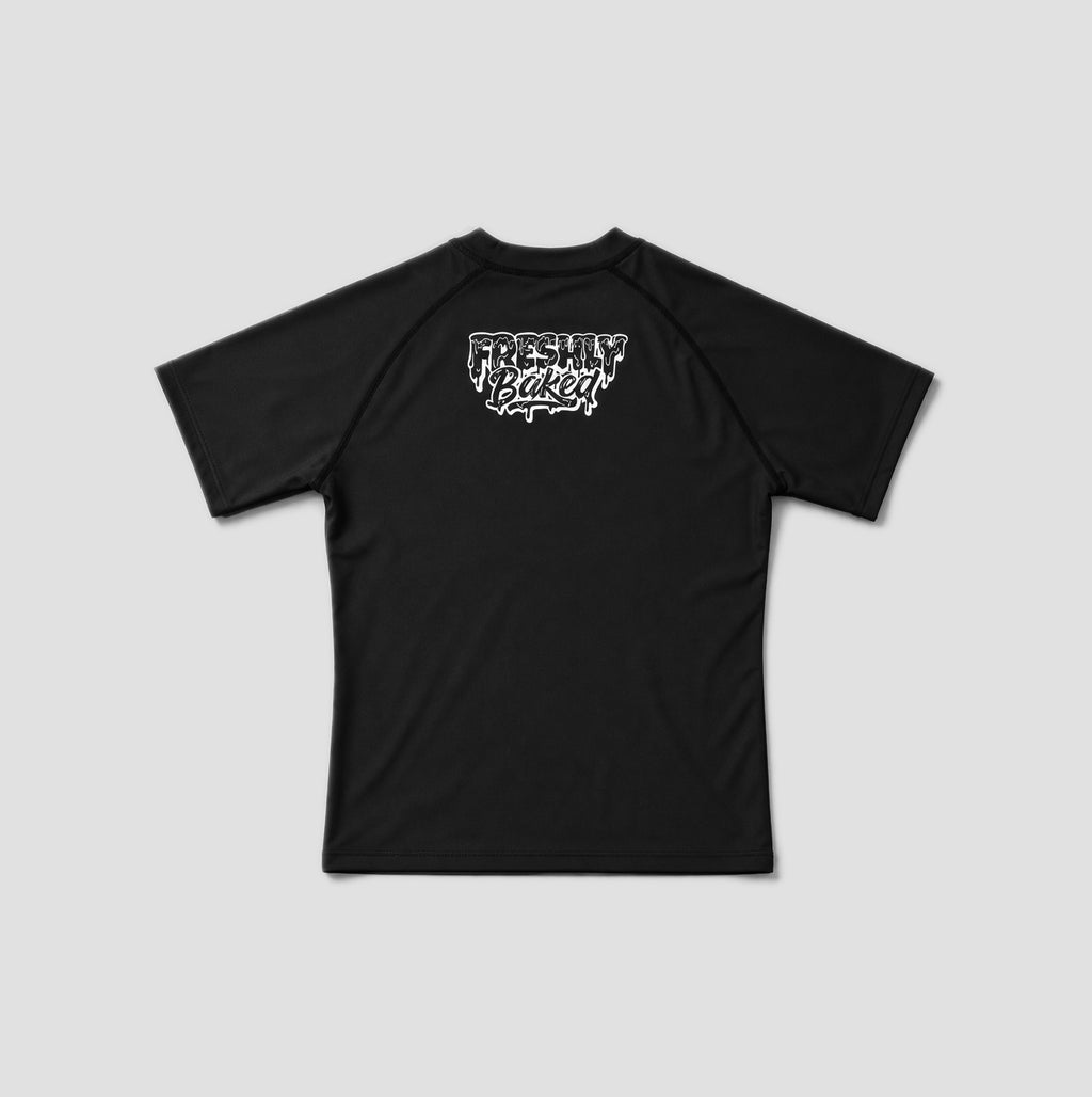 A black short-sleeve rash guard with raglan sleeves and a white 'Freshly Baked' logo printed on the chest, displayed against a solid gray background.