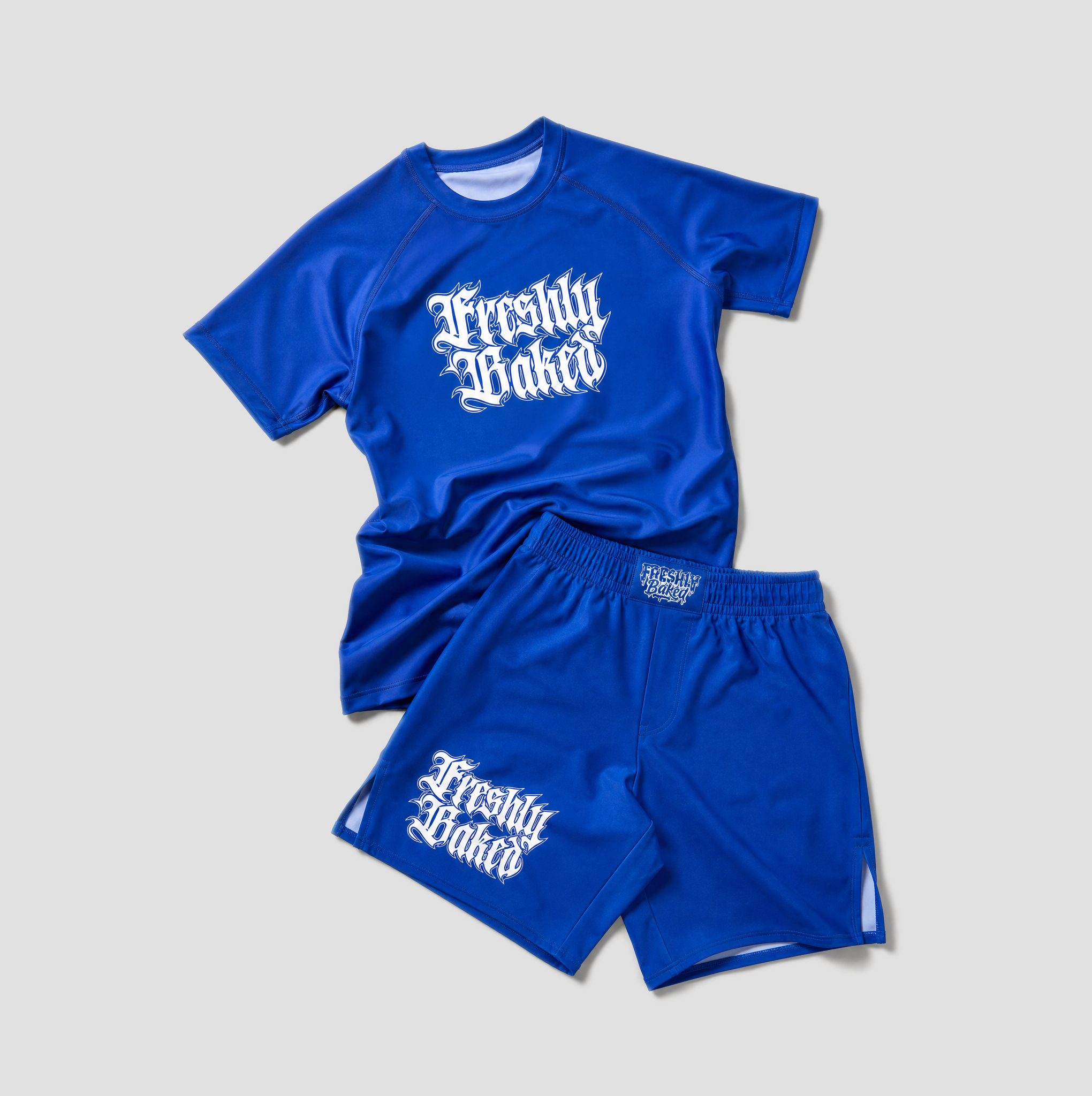 A two-piece athletic set consisting of a royal blue short-sleeve raglan t-shirt and matching shorts, both featuring the text 'Freshly Baked' printed in a white gothic font. The t-shirt is laid flat on top of the shorts against a neutral light gray background.