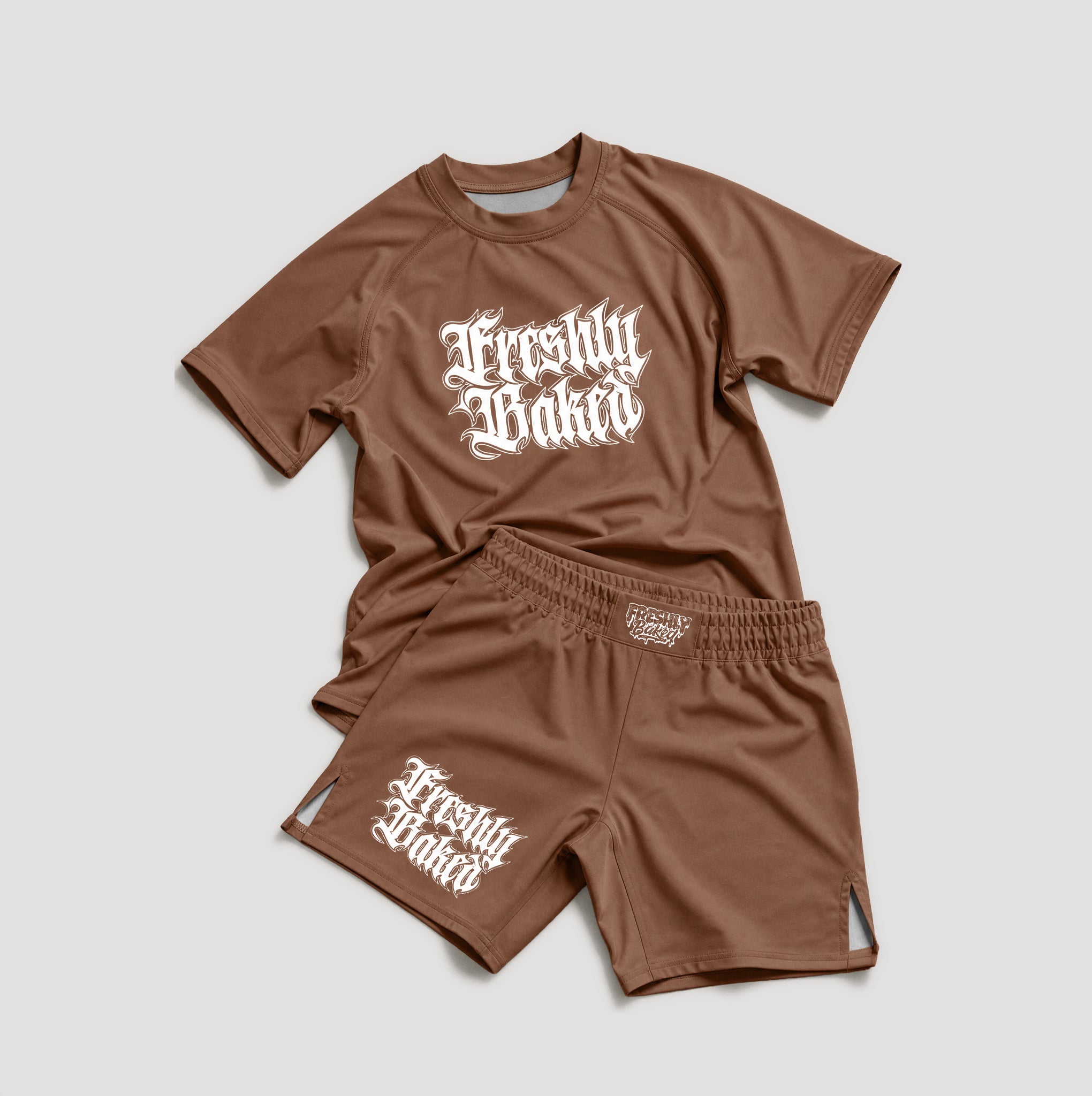 A two-piece kids' athletic set consisting of a brown short-sleeve t-shirt and matching shorts, both featuring a white Gothic-style 'Freshly Baked' graphic on the front and side. The set is laid flat on a light gray background.