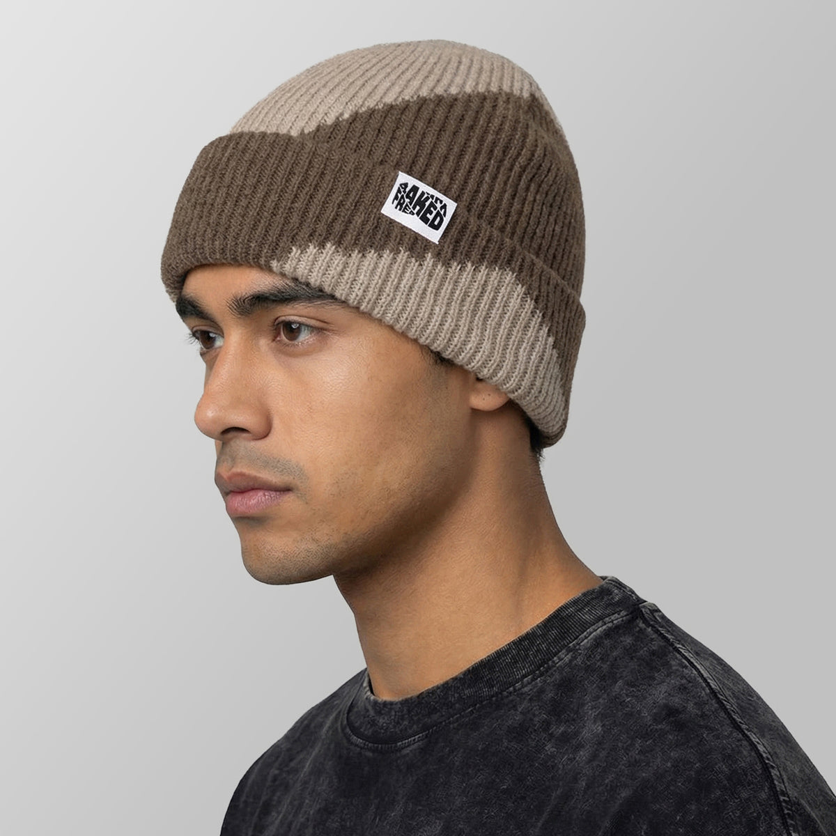 A man in profile wearing a ribbed knit beanie hat with horizontal brown and beige stripes and a small white brand patch that reads FRESHLY BAKED
