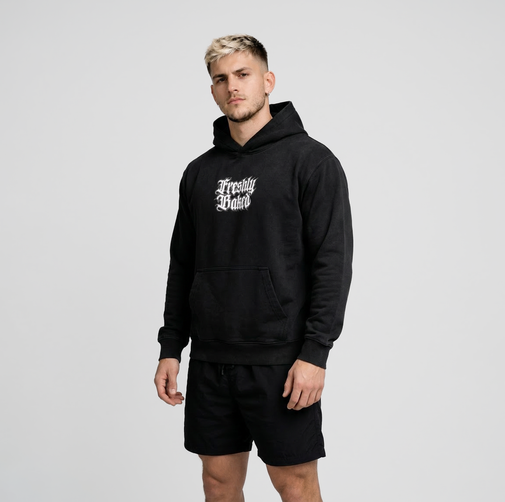 A man wearing a black pullover hoodie with 'Freshly Baked' printed in white gothic lettering on the chest, paired with black shorts, against a light gray studio background.