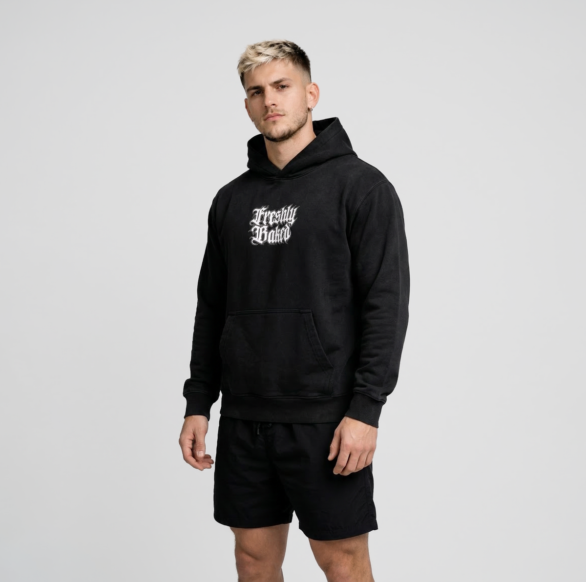 A man wearing a black pullover hoodie with 'Freshly Baked' printed in white gothic lettering on the chest, paired with black shorts, against a light gray studio background.