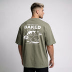 A man wearing an oversized olive green t-shirt, viewed from the back. The shirt features a large white graphic print on the back with the text 'FRESHLY BAKED', 'NUGS', and 'EST.