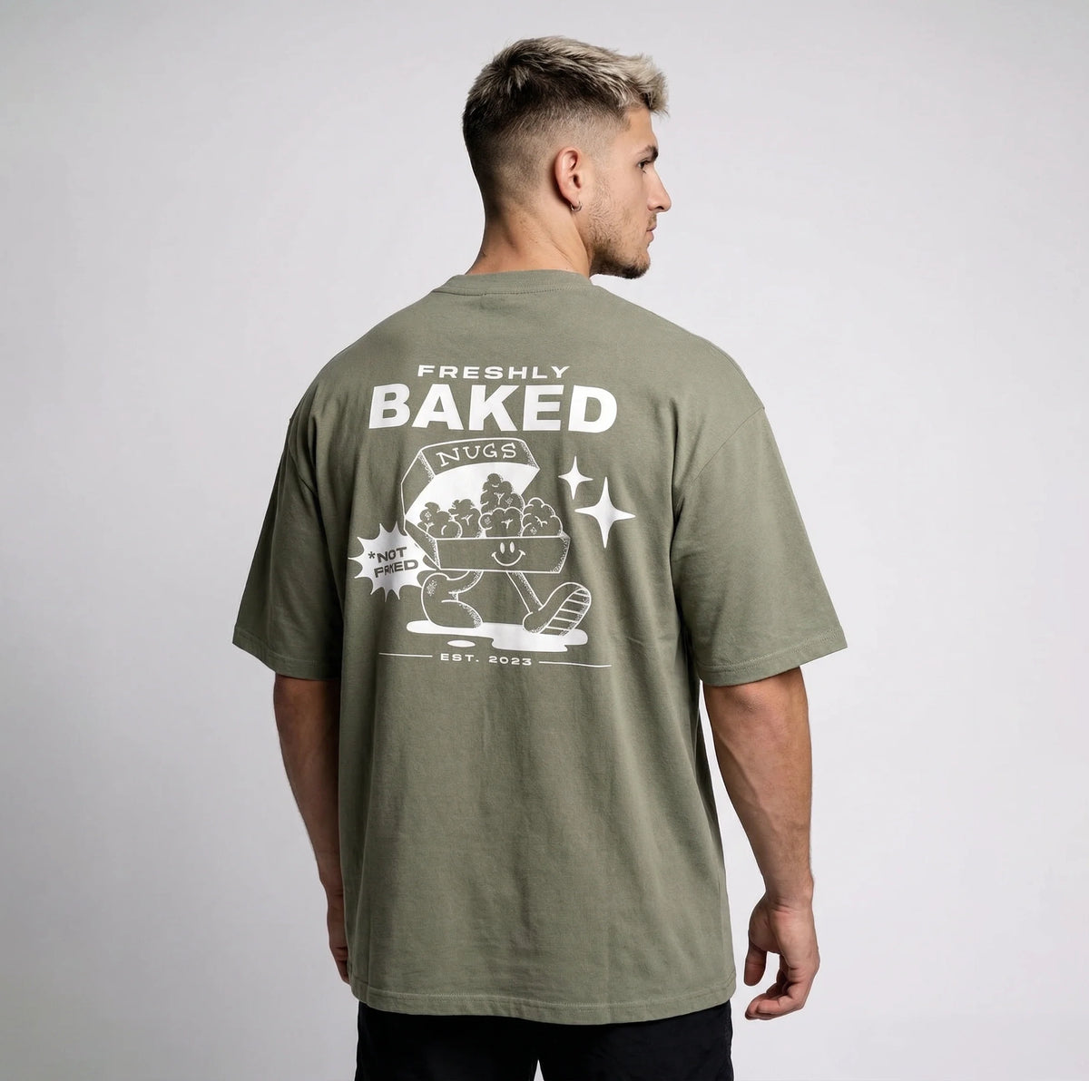 A man wearing an oversized olive green t-shirt, viewed from the back. The shirt features a large white graphic print on the back with the text 'FRESHLY BAKED', 'NUGS', and 'EST.