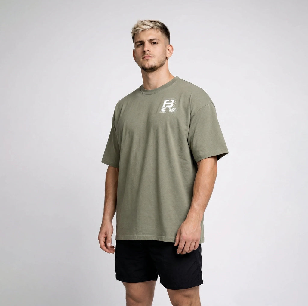 A man wearing an oversized sage green t-shirt with a small white graphic logo on the left chest, paired with black athletic shorts, standing against a light gray studio background.