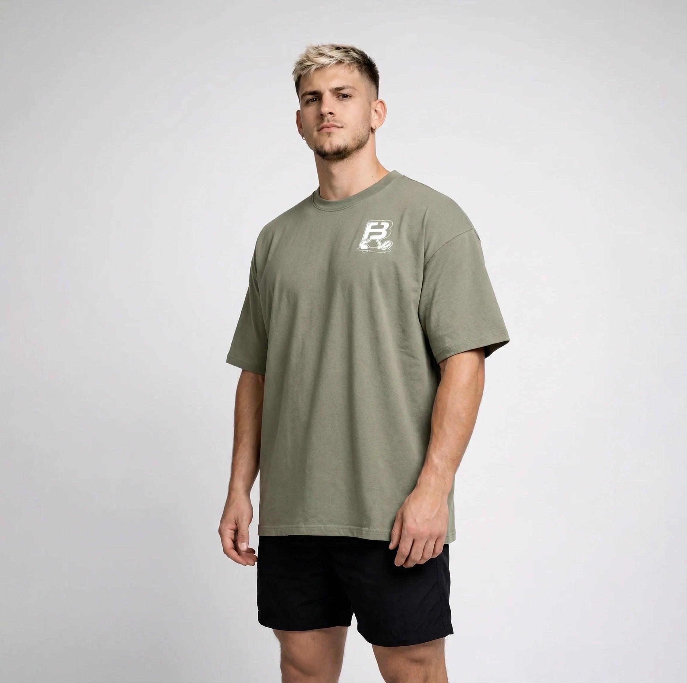 A man wearing an oversized sage green t-shirt with a small white graphic logo on the left chest, paired with black athletic shorts, standing against a light gray studio background.