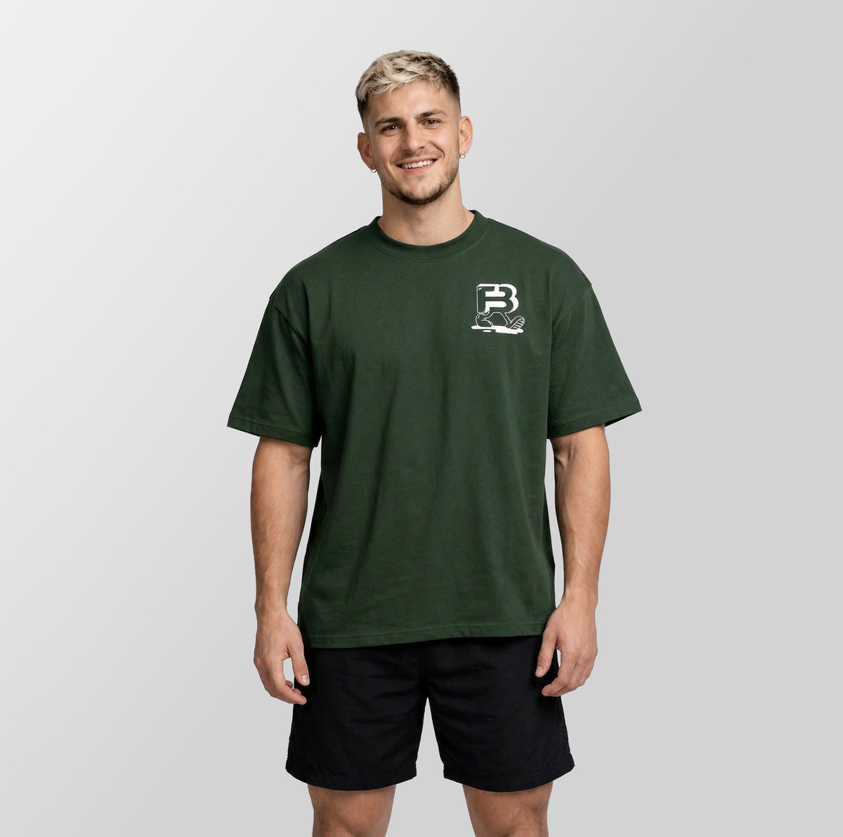 A man wearing a dark forest green crewneck t-shirt with a small white graphic logo on the left chest, paired with black athletic shorts, standing against a light gray studio background.
