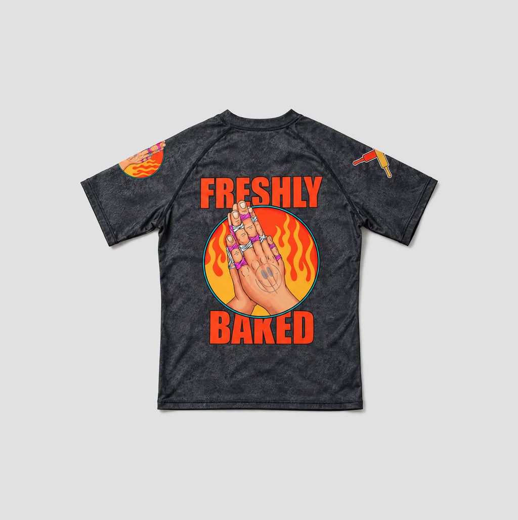 A dark gray acid-wash short-sleeve t-shirt with a graphic on the back featuring a hand with rings, flames, and the text 'FRESHLY BAKED' in orange, shown on a light gray background.