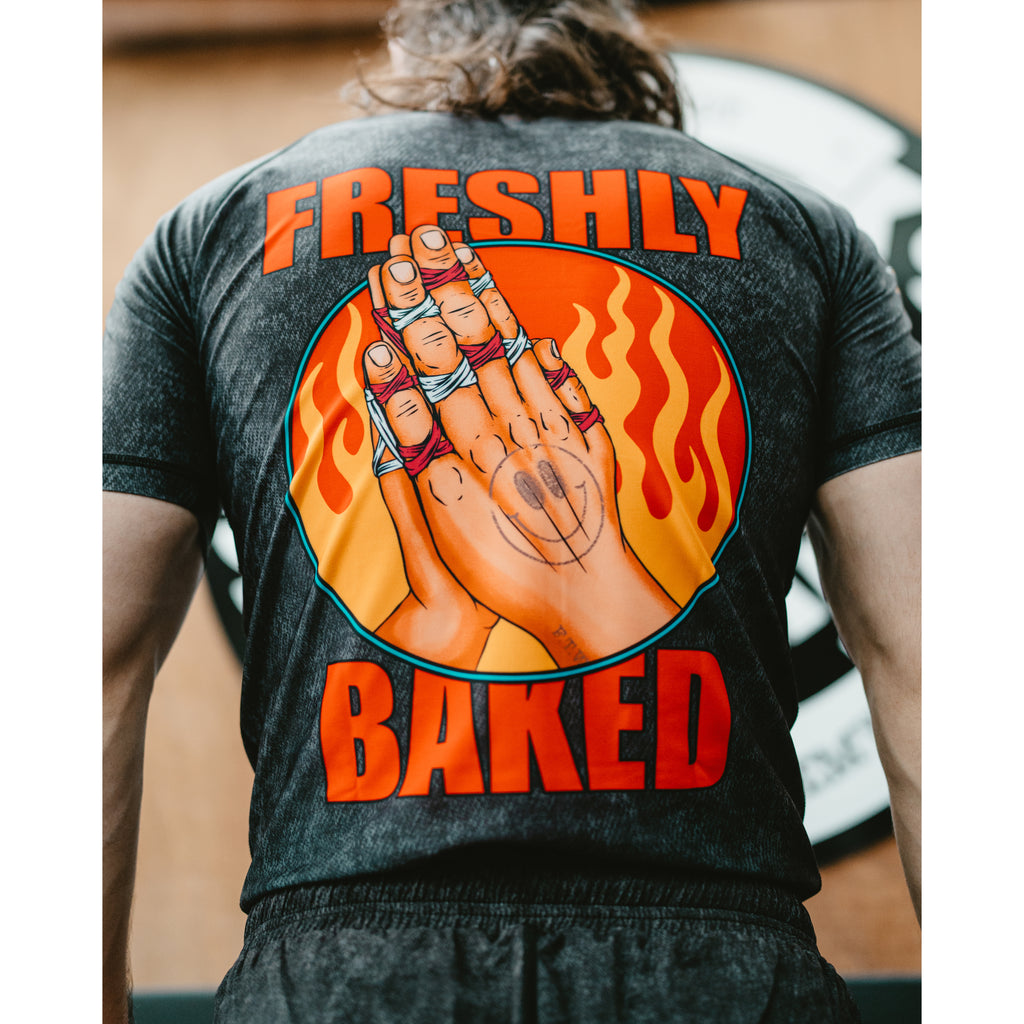 A person viewed from behind wearing a dark charcoal gray t-shirt with a large graphic on the back. The graphic features the words 'FRESHLY BAKED' in bold orange letters surrounding an illustration of hands with rings inside a circular flame design.