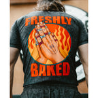 A person viewed from behind wearing a dark charcoal gray t-shirt with a large graphic on the back. The graphic features the words 'FRESHLY BAKED' in bold orange letters surrounding an illustration of hands with rings inside a circular flame design.