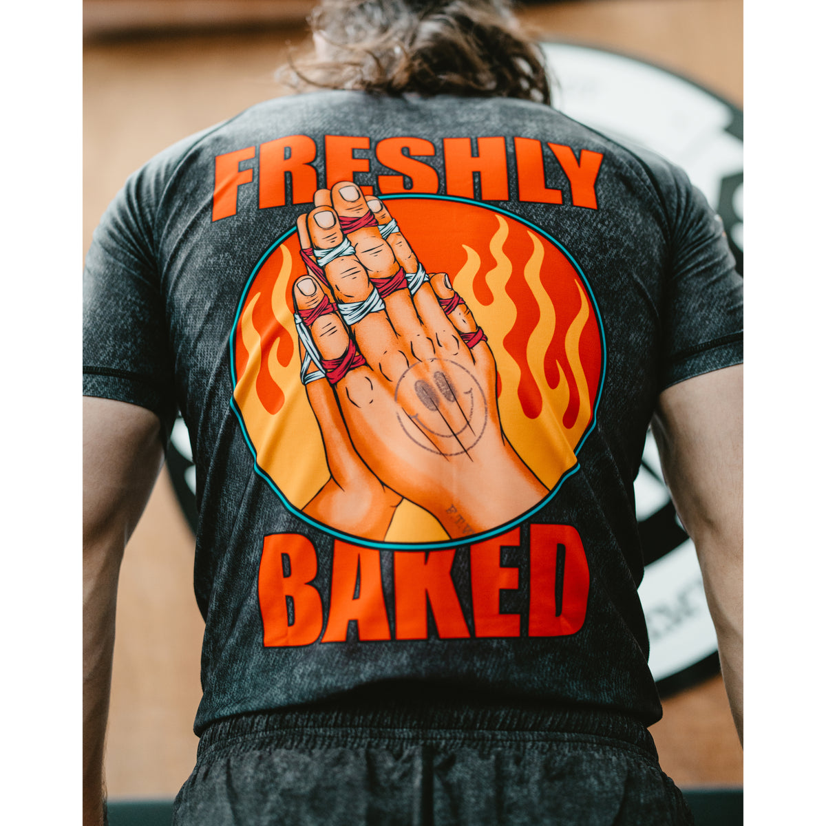 A person viewed from behind wearing a dark charcoal gray t-shirt with a large graphic on the back. The graphic features the words 'FRESHLY BAKED' in bold orange letters surrounding an illustration of hands with rings inside a circular flame design.